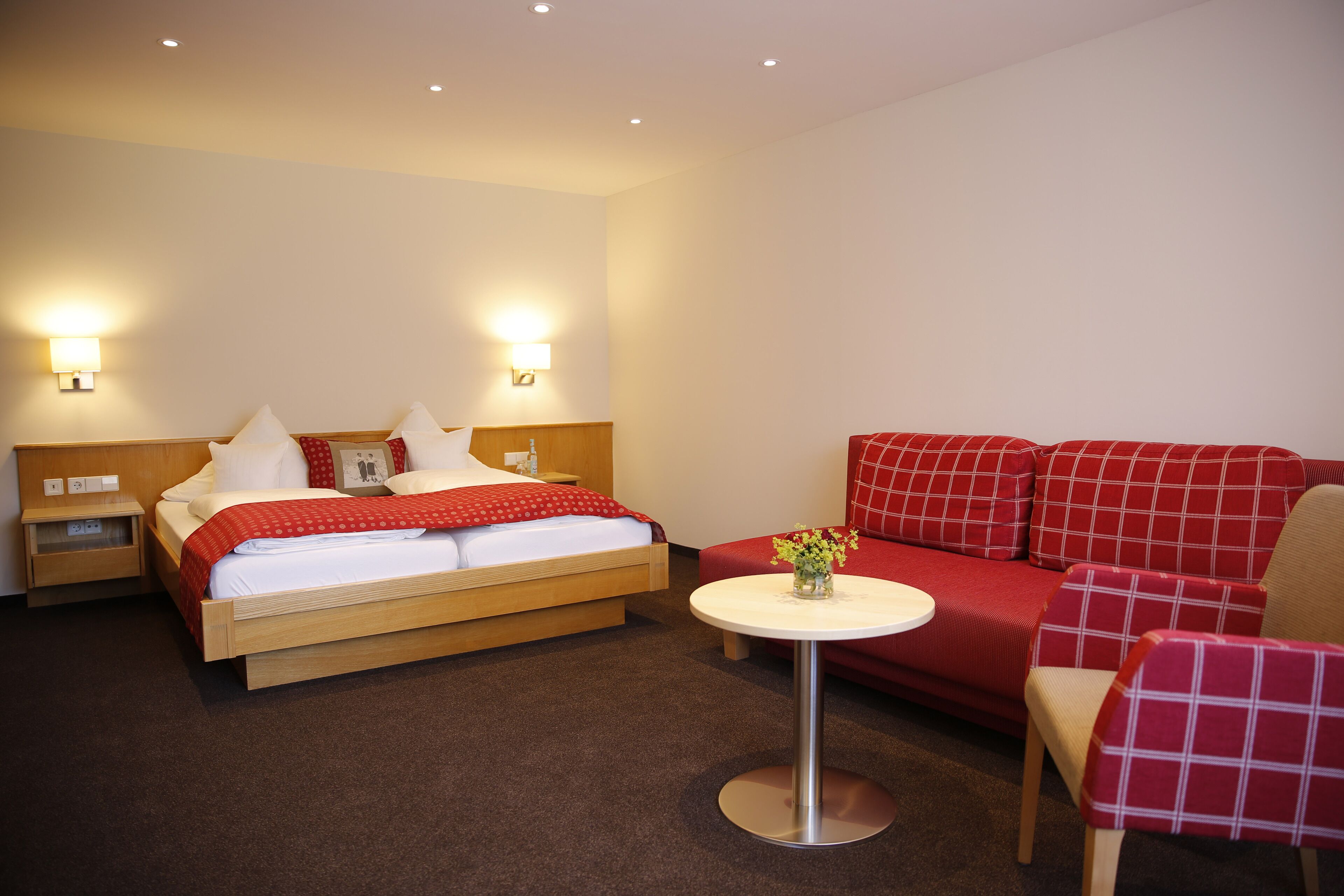 A hotel room with a bed, bedside tables, a small round table with a vase of flowers, and a red sofa with matching armchairs.