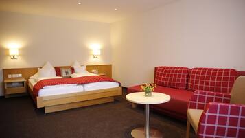 A hotel room with a bed, bedside tables, a small round table with a vase of flowers, and a red sofa with matching armchairs.