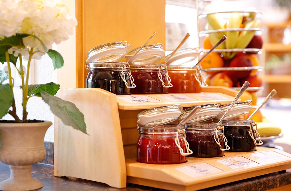 A display of various jams with labels, a potted plant, and a fruit basket in the background.