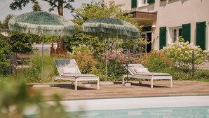 A poolside area with two lounge chairs and a thatched umbrella, surrounded by lush greenery and a building with green shutters. - Hotel Restaurant Mühle (Binzen)