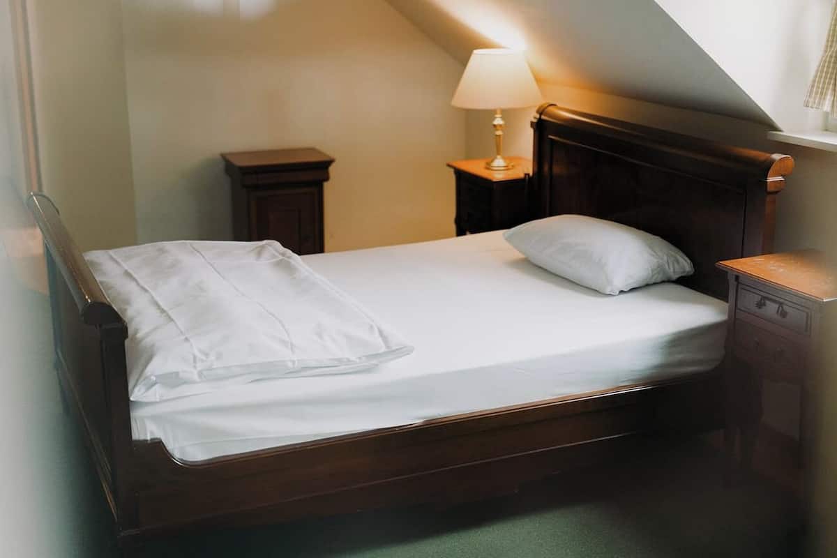 A single bed with a wooden frame, a white mattress, and a white pillow. There is a nightstand with a lamp on it to the right of the bed.
