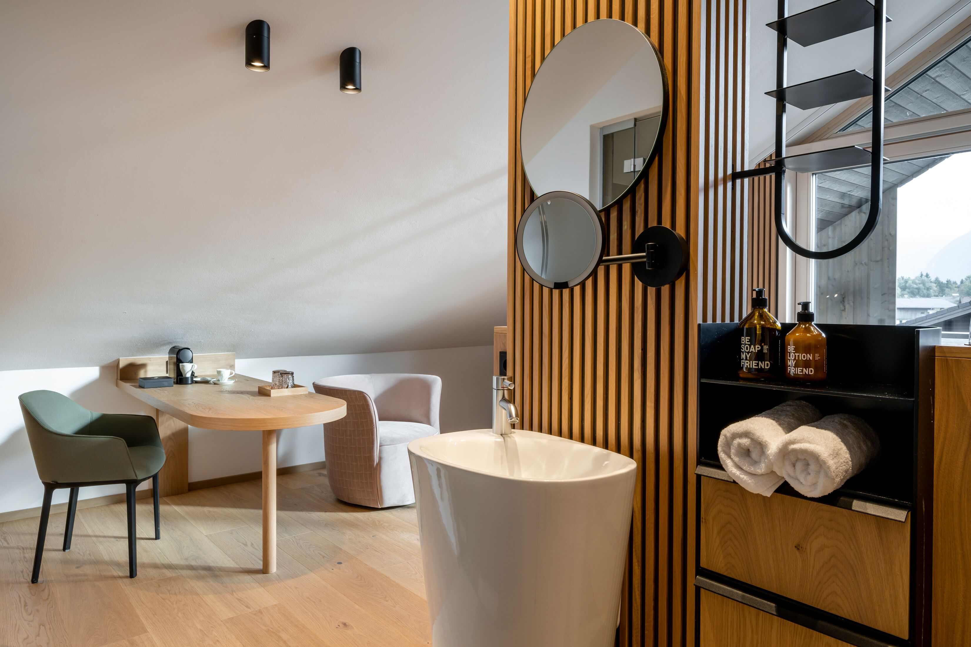 A modern bathroom with a wooden vanity, a round mirror, a sink, and a shelf with towels and toiletries.