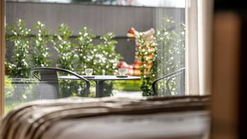 A view from a window of a room with a bed, a small table with a cup and saucer, and a view of a garden with a fence and a statue.