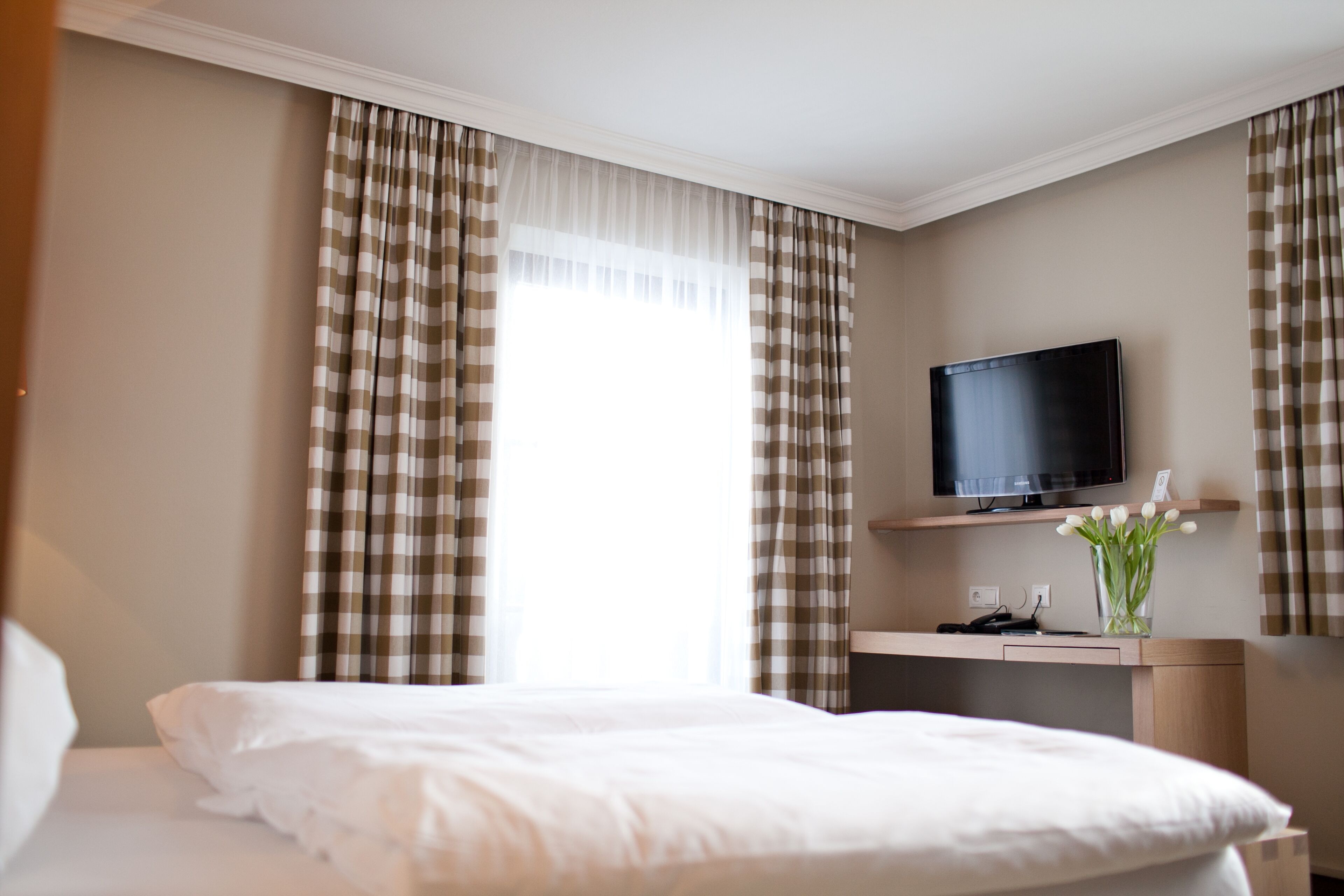 Una habitación de hotel con una cama, un televisor sobre un soporte, un escritorio con un florero y cortinas a cuadros.