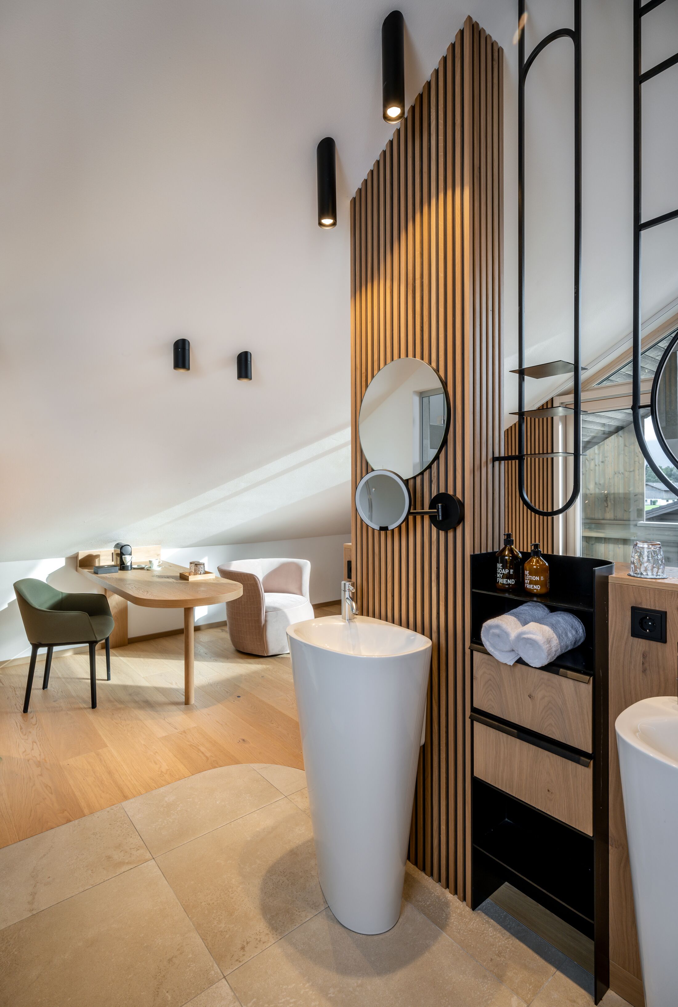 Un baño moderno con un lavamanos grande de color blanco, un espejo redondo, paneles de madera y una pequeña zona de estar con una mesa y sillas.