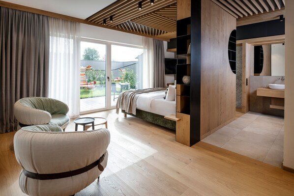 A modern bedroom with a large bed, a seating area with chairs, a small table, and a bathroom with a sink and mirror. - NEUWIRT – Hotel & Wirtshaus (Bad Vigaun)