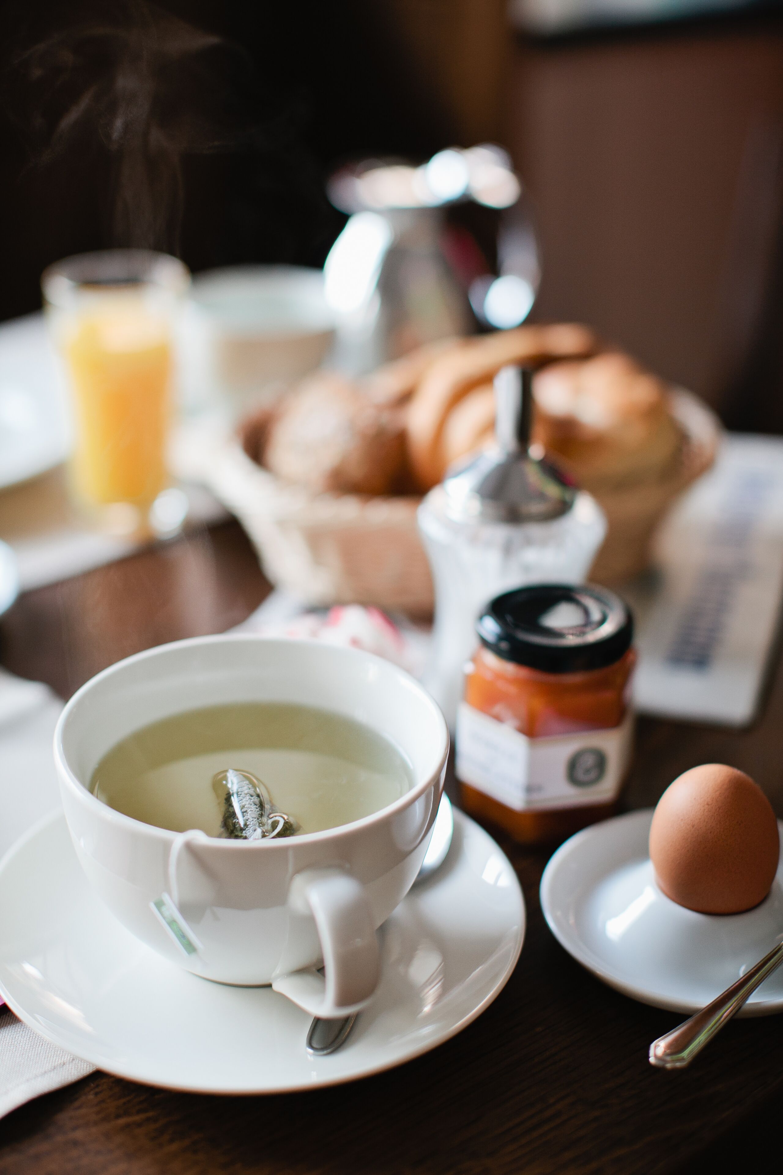 Et morgenbord med en kop te, et glas appelsinjuice, en croissant, et syltetøjsglas og et hårdkogt æg på et bord.