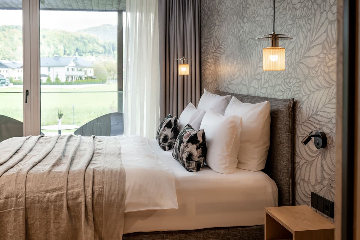 A neatly made bed with a beige blanket and white pillows, a wooden bedside table, and a view of a green landscape through a window.