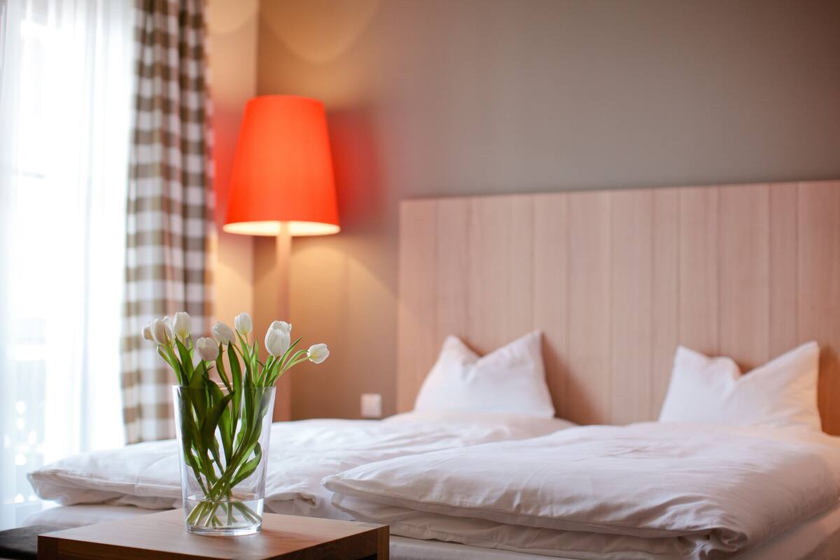 A hotel room with a bed, a bedside table with a vase of flowers, and a red lamp.