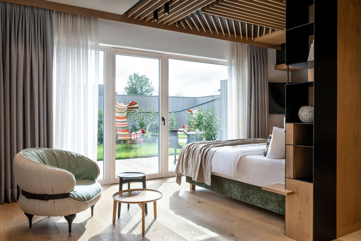 A modern bedroom with a bed, a green armchair, a small table, and a sliding glass door leading to a backyard.
