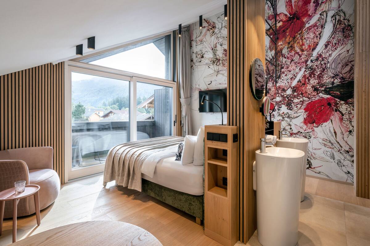 A modern bedroom with a large window, a bed, a bedside table, a chair, and a bathroom with a large sink.