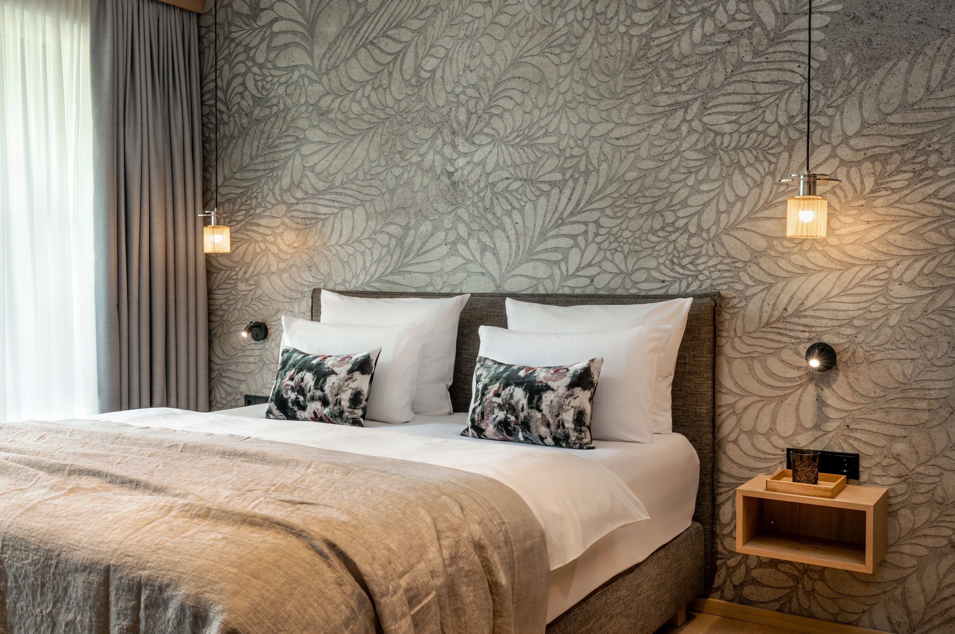 A neatly made bed with white and patterned pillows, a wooden nightstand with a small shelf, and a wall with a leaf-patterned wallpaper.