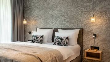 A neatly made bed with white and patterned pillows, a wooden nightstand with a small shelf, and a wall with a leaf-patterned wallpaper.