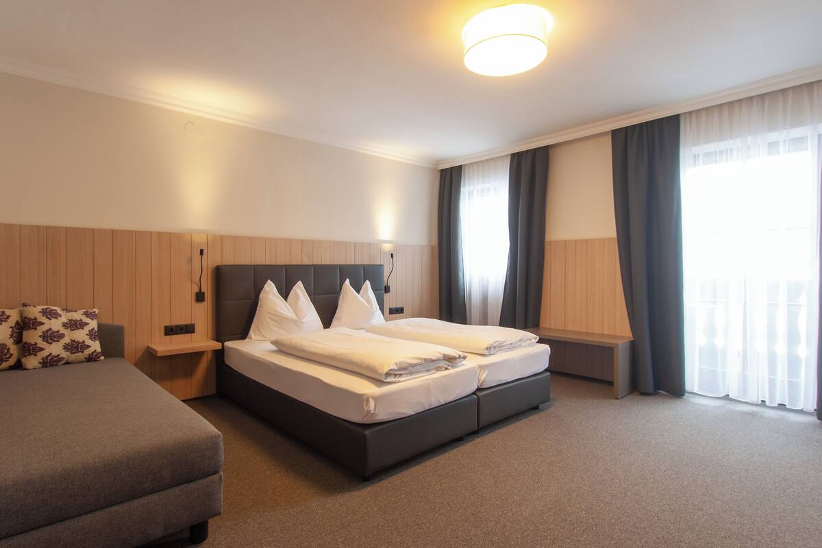A hotel room with a double bed, a sofa, a nightstand, a lamp, and a large window with curtains.