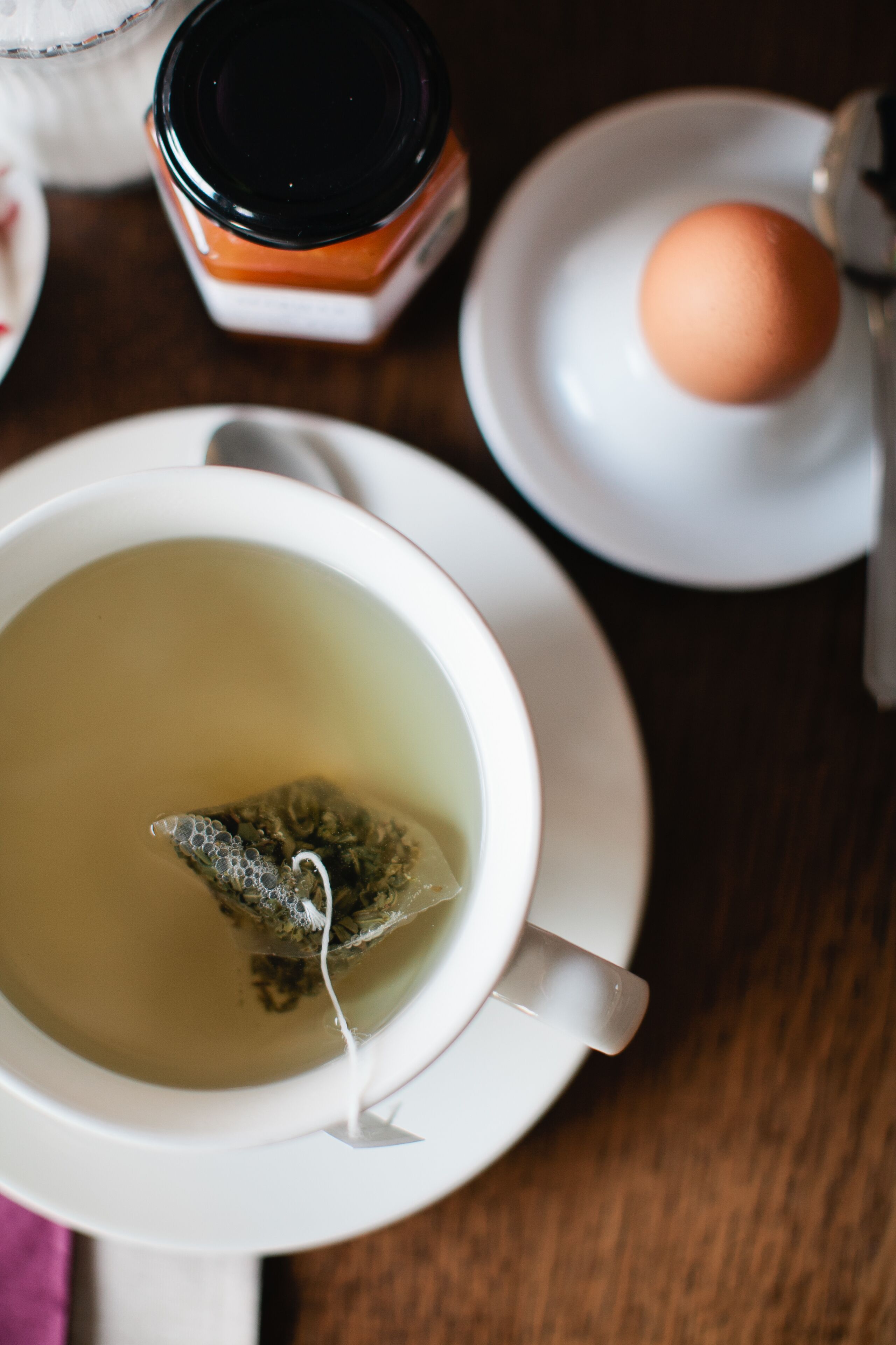 Una taza de té con una bolsita, un plato con un huevo y un frasco de mermelada sobre una mesa.