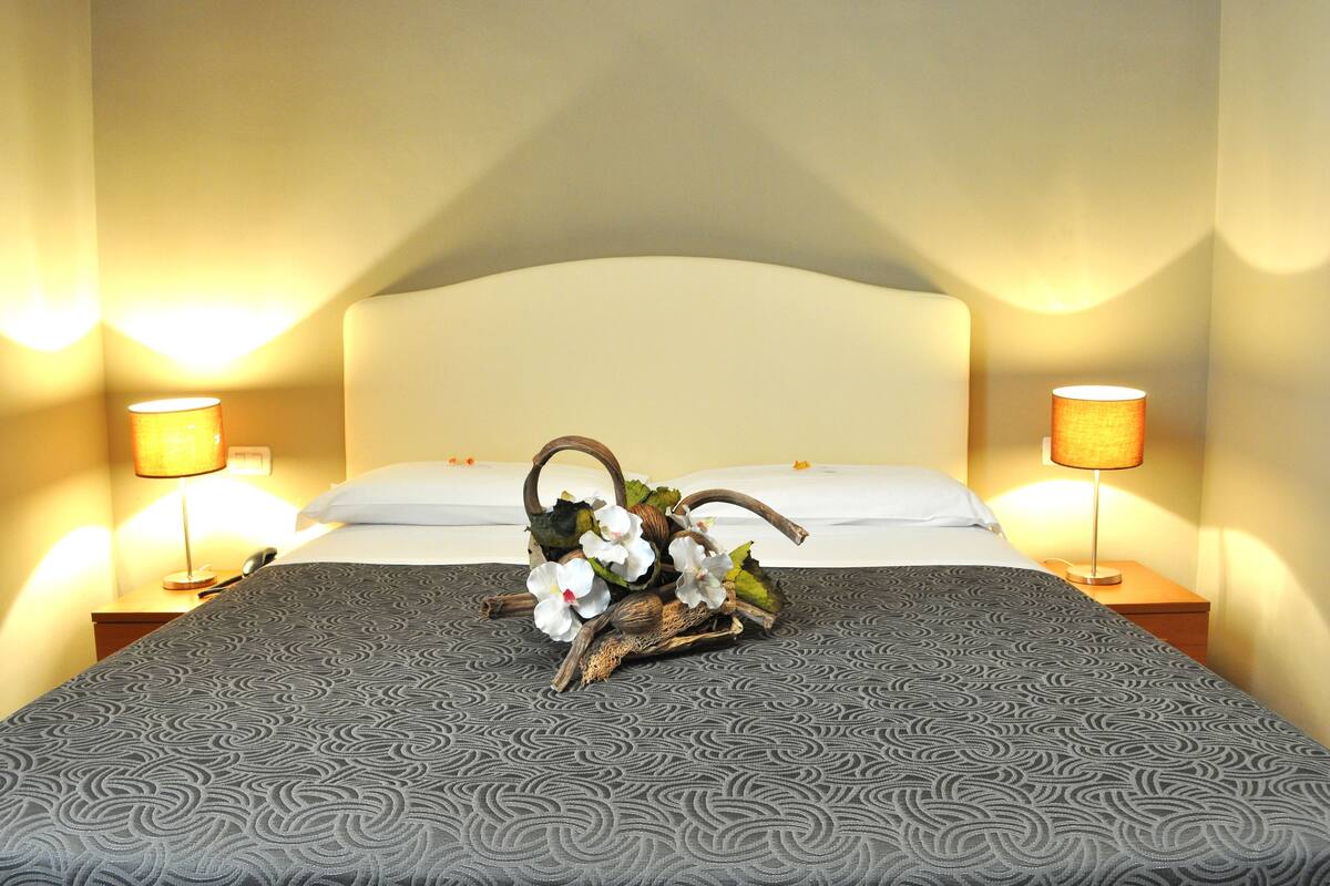 Een netjes opgemaakt bed met een bedsprei met dessin, een decoratieve bloemenboeket en twee bedlampjes.