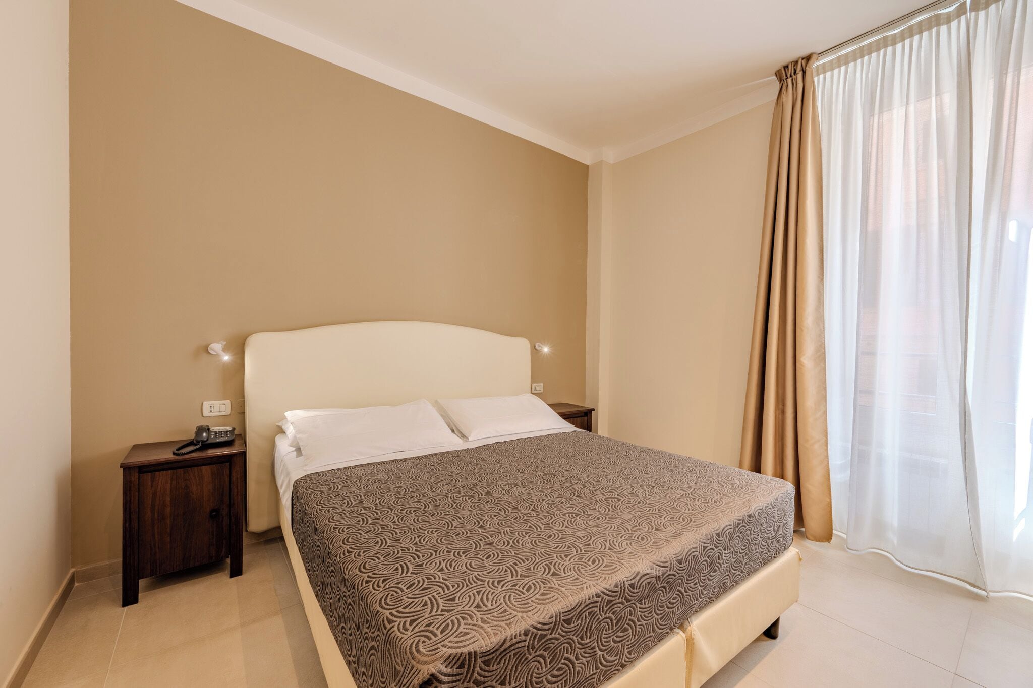 Ein ordentlich eingerichtetes Schlafzimmer mit einem großen Bett, einem Nachttisch mit Telefon und einem Fenster mit transparenten Vorhängen.