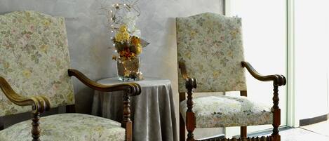 Two upholstered chairs with floral patterns, a small table with a vase of flowers, and a plain wall with a window.