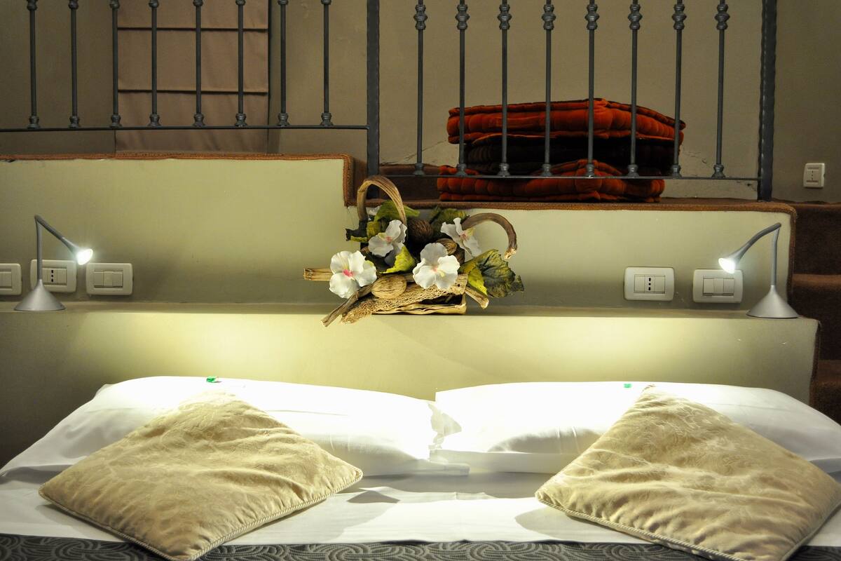 Een opgemaakt bed met twee kussens, een bedlampje en een decoratieve mand met bloemen op een wandplank.