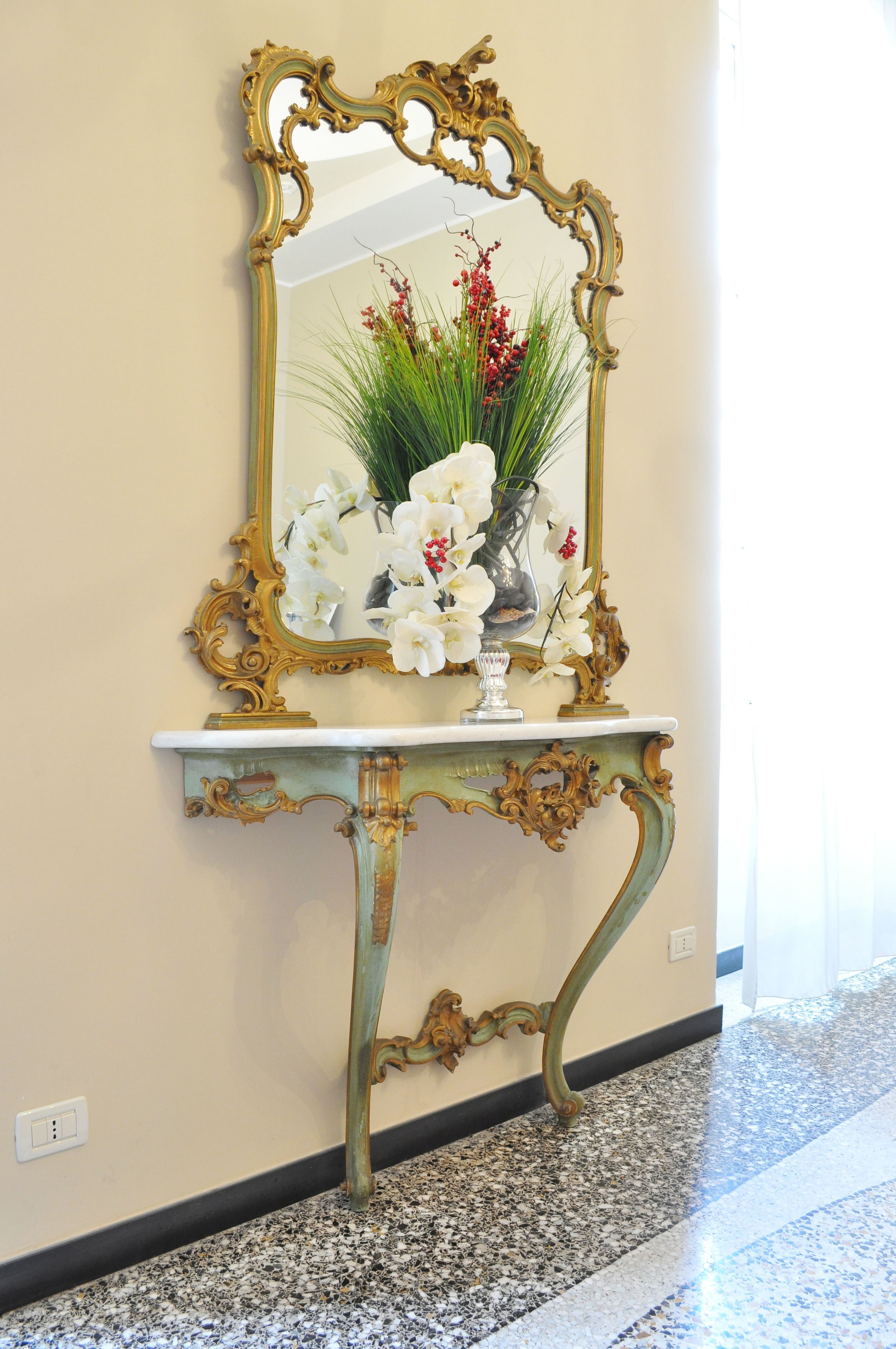 Un miroir avec un cadre doré orné, un vase blanc rempli de fleurs et une console décorative adossée à un mur.