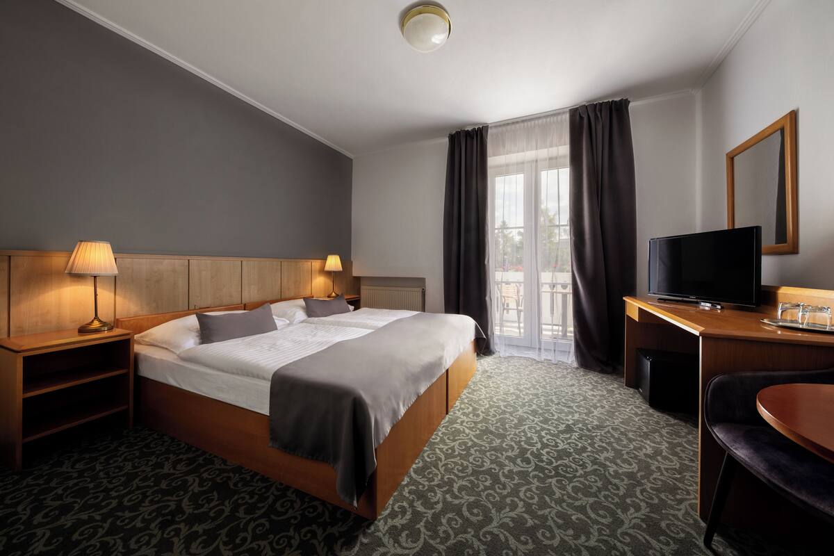 Habitación de hotel con cama, mesitas de noche, un escritorio con televisor y un balcón con cortinas.