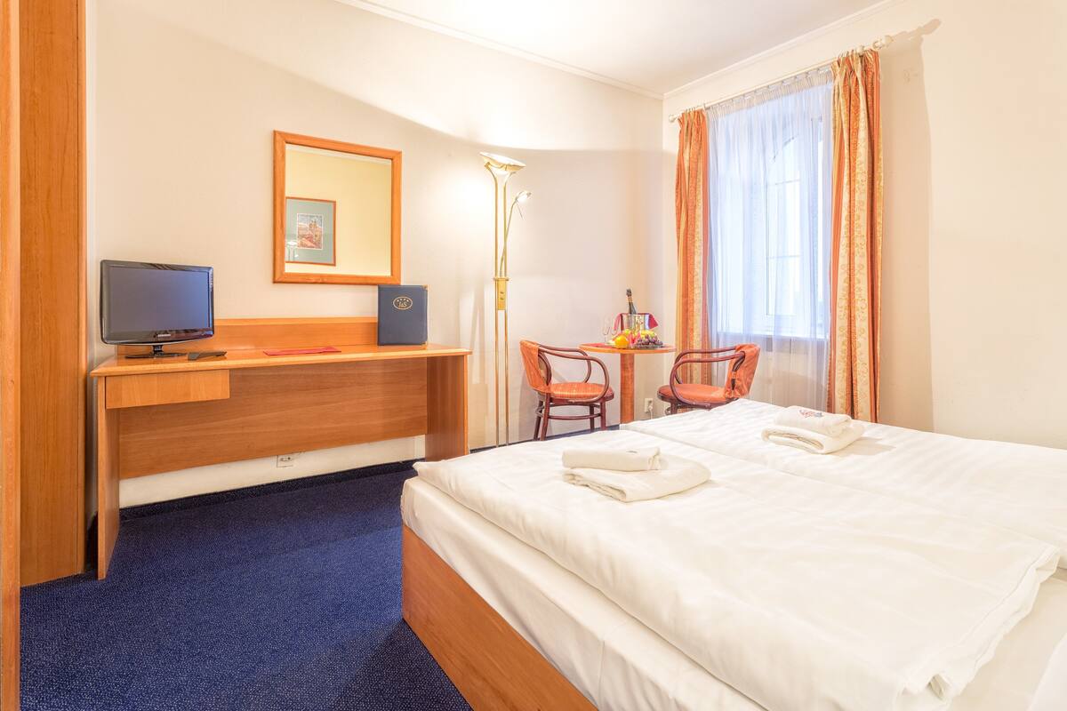 Habitación de hotel con cama, escritorio, computadora, silla, lámpara y ventana con cortinas.