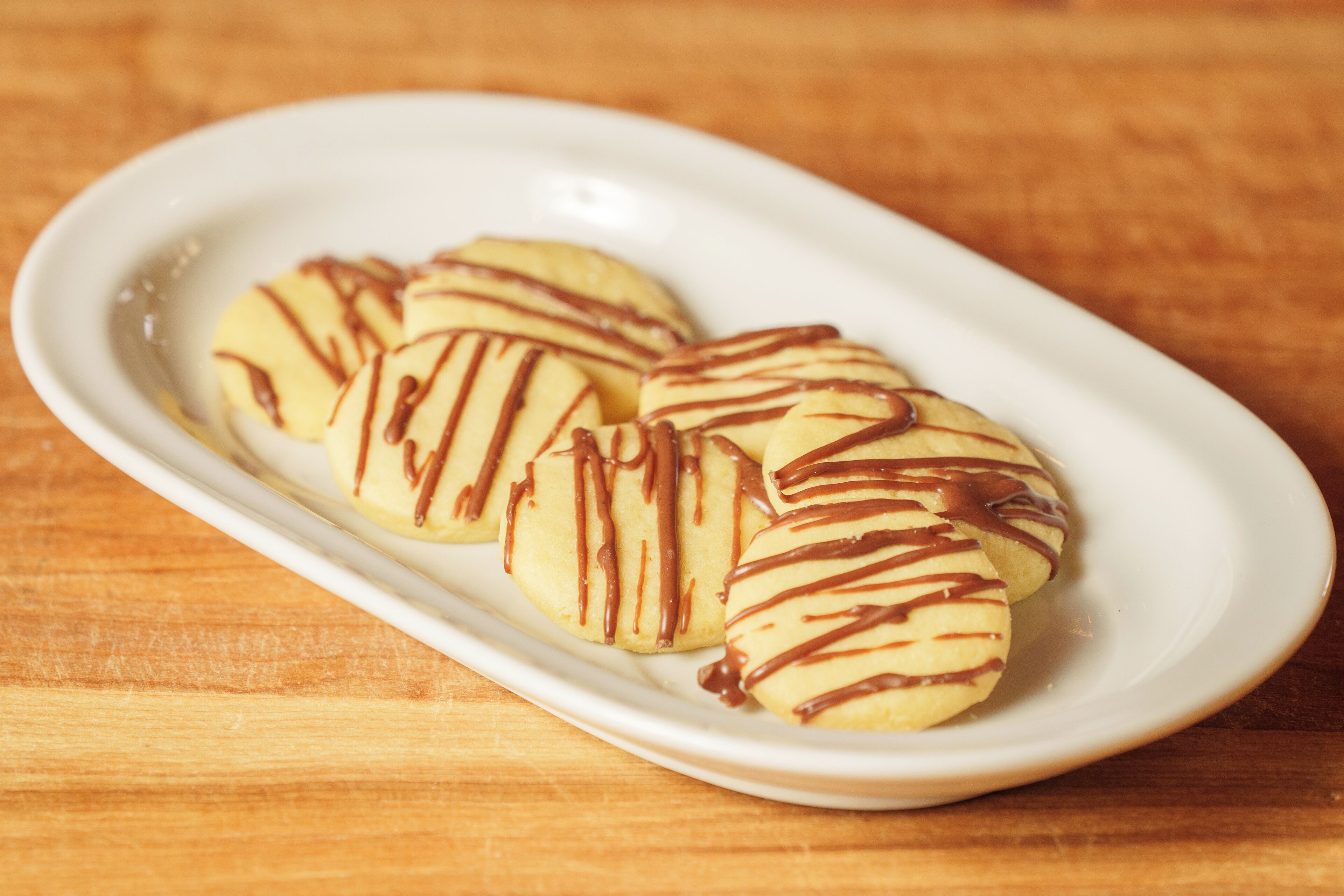 A white oval plate with four round cookies drizzled with chocolate on a wooden surface.