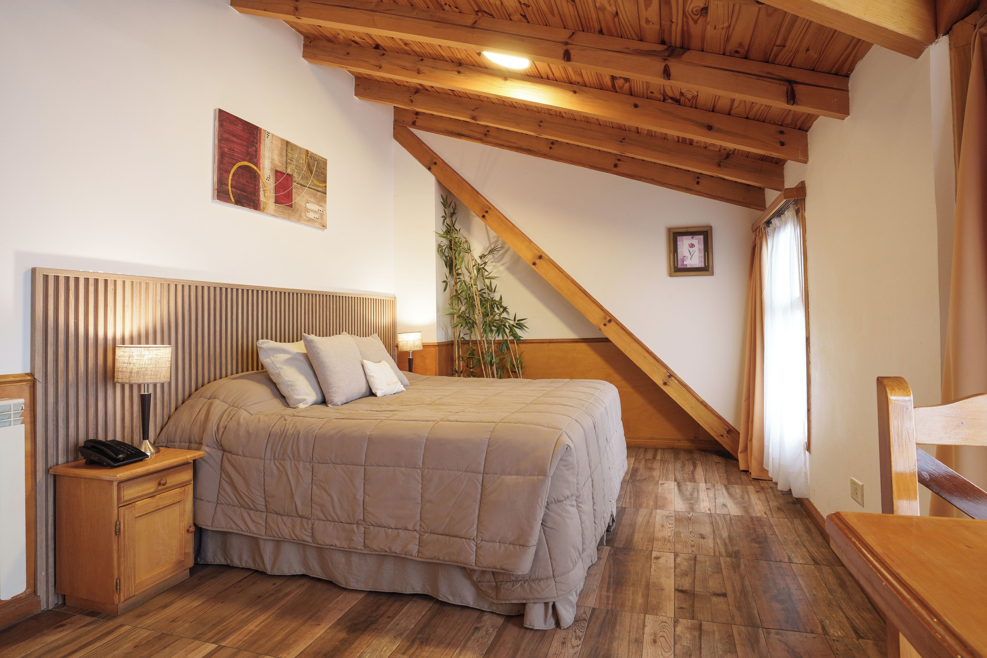 A bedroom with a wooden headboard, a bed with a beige quilt, a bedside table with a lamp, and a wooden staircase.