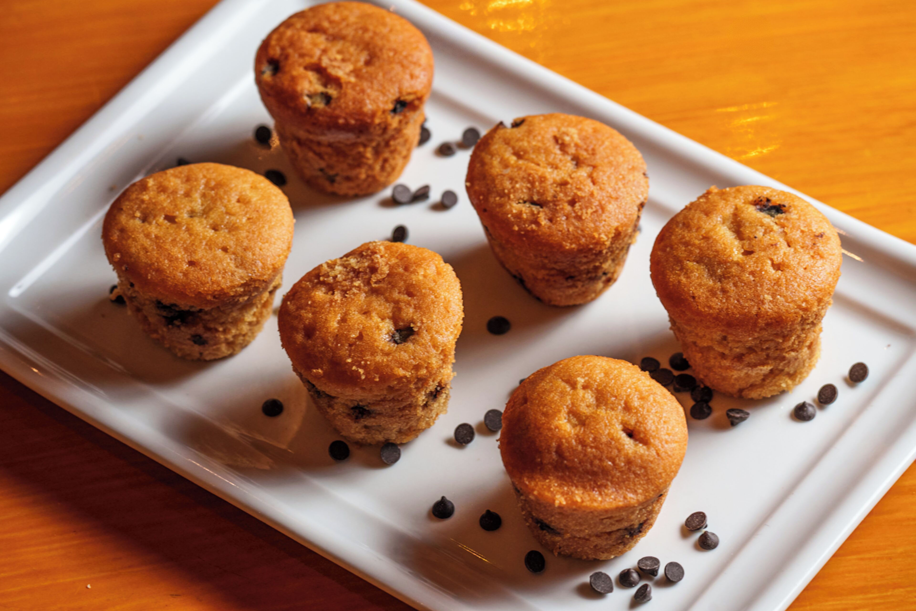A white rectangular plate with six muffins and chocolate chips on a wooden table.