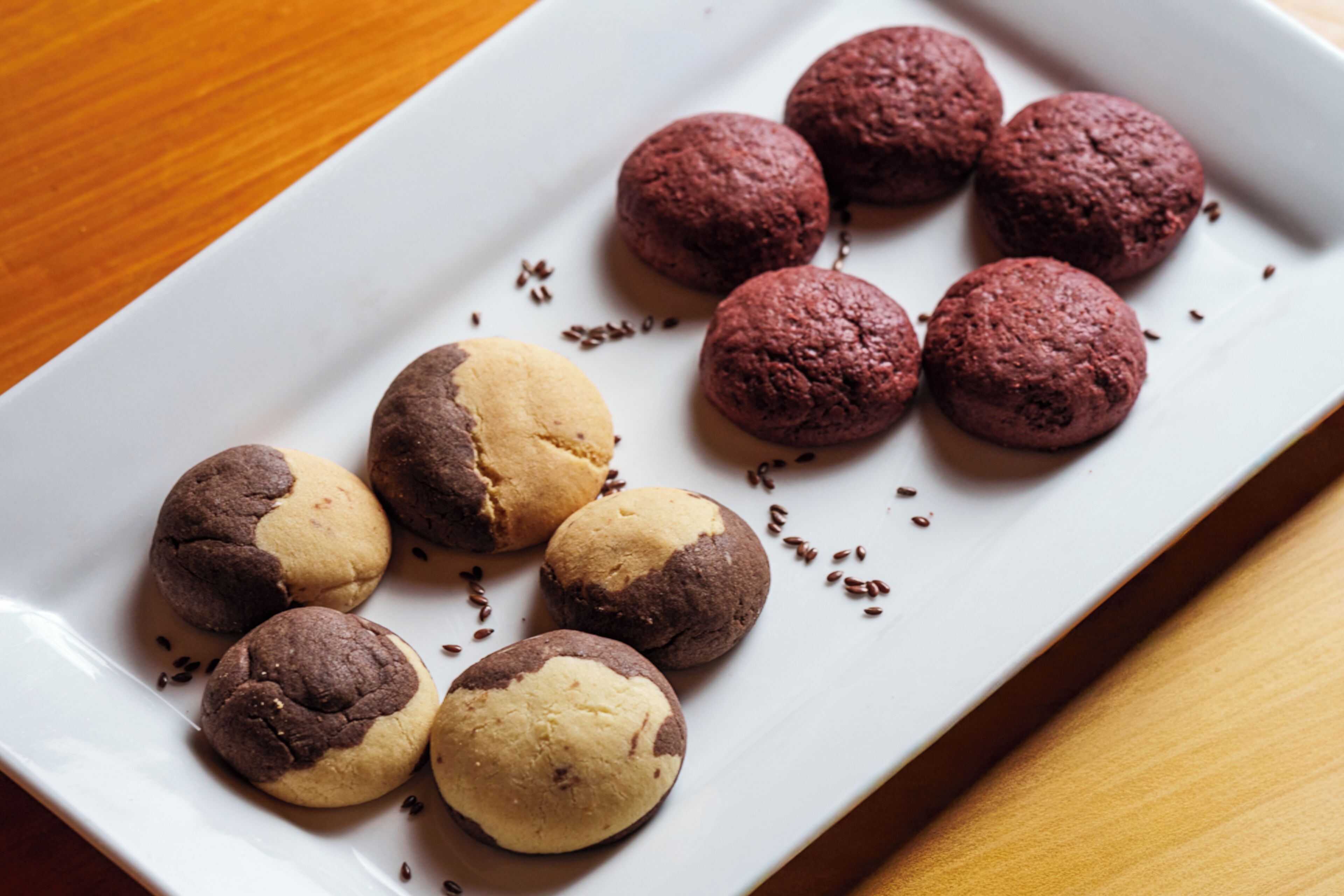 A white rectangular plate with a variety of cookies, some with chocolate chips and others with a chocolate and vanilla swirl, on a wooden table.