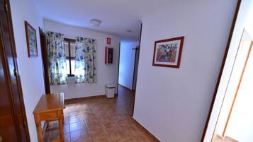 Un pasillo con una mesa de madera, una ventana con cortinas florales, un extintor y un cuadro en la pared.