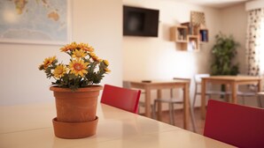 Une table à manger surmontée d’un pot en terre cuite rempli de fleurs jaunes, des chaises rouges et une carte accrochée au mur.