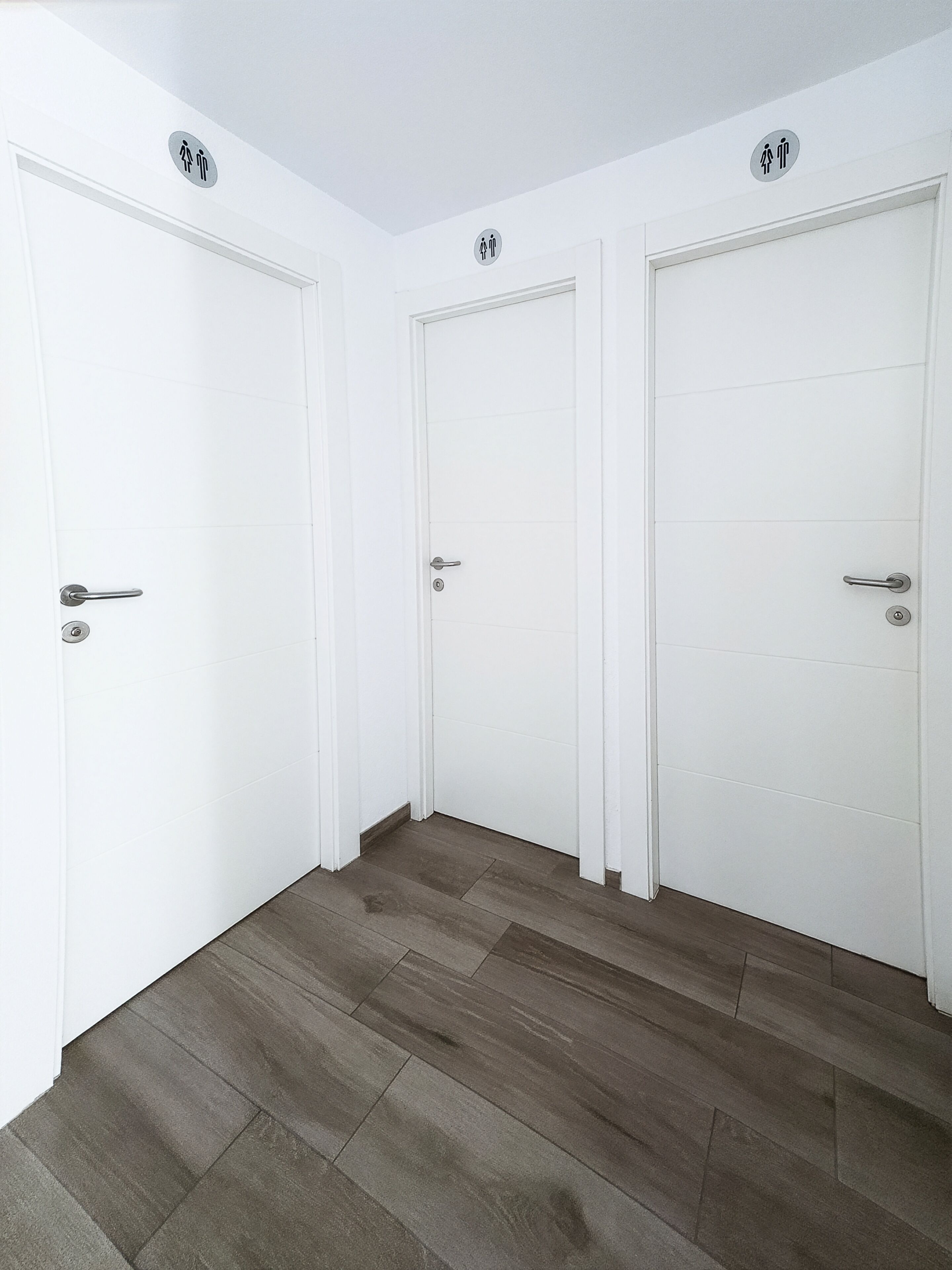 Um corredor com três portas brancas, cada uma com maçaneta e fechadura. O chão é revestido de azulejos cinzas.
