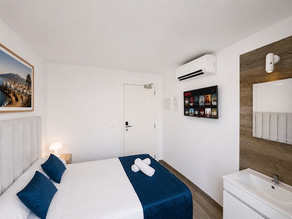A hotel room with a bed, a wall-mounted TV, a bathroom with a sink, and an air conditioning unit.