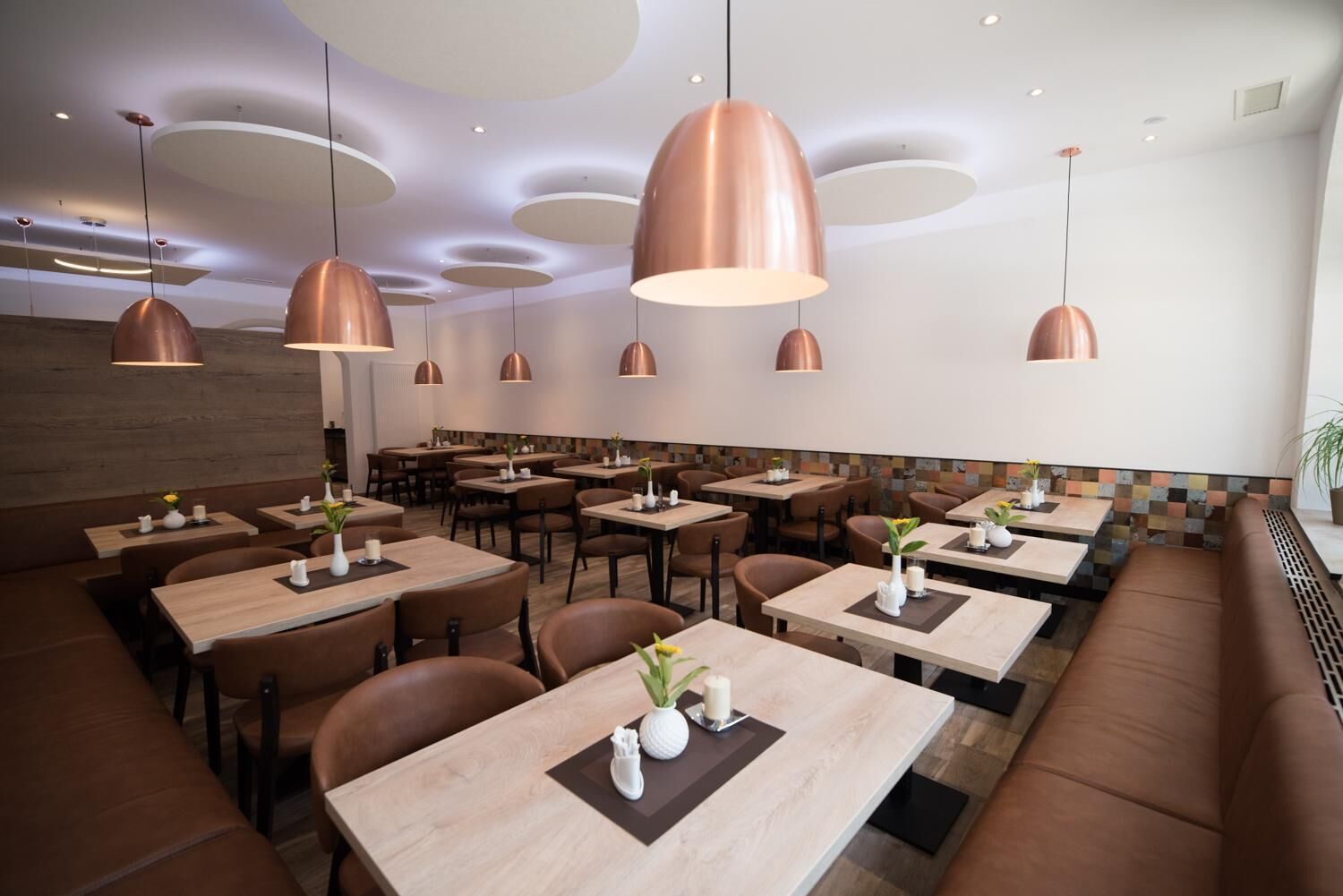 A modern restaurant with wooden tables and brown leather booths, featuring copper pendant lighting and a patterned tile backsplash.