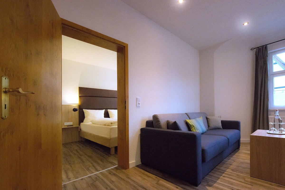 A hotel room with a wooden door, a bed with white bedding, a dark-colored sofa with cushions, and a small table with a bottle and glasses.