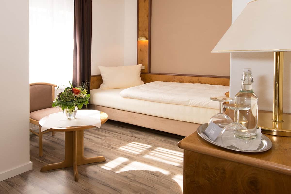 A hotel room with a bed, a small table with a vase of flowers, a chair, a bedside table with a lamp and glassware.