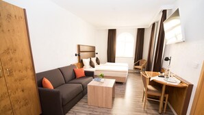 Una habitación de hotel moderna con un sofá gris oscuro, una cama blanca, un escritorio de madera y un armario.