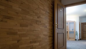 Un pasillo con suelo de madera y pared con motivo de espiga. Una puerta de madera con manija de latón está abierta, mostrando una habitación con un armario y un florero con flores.