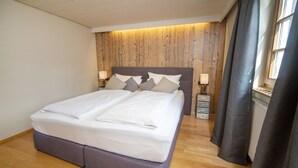 Ein ordentlich bezogenes Bett mit einem Holzkopfstück, ein Nachttisch mit Lampe und ein Fenster mit Vorhängen.