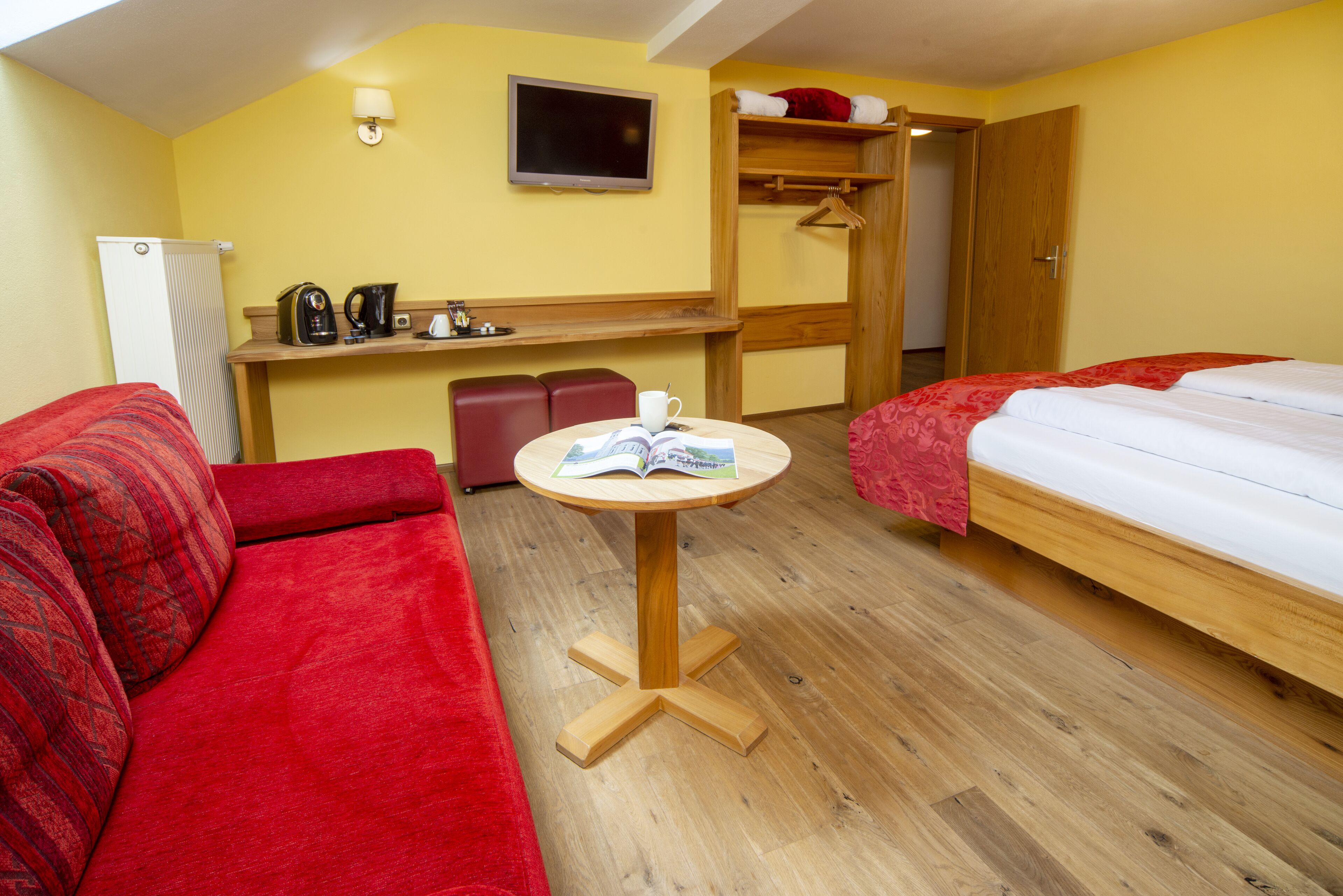 Ein Hotelzimmer mit rotem Sofa, einem Bett, einem kleinen runden Tisch mit einem Buch, einem Fernseher, einer Minibar und einem Holzschrank.