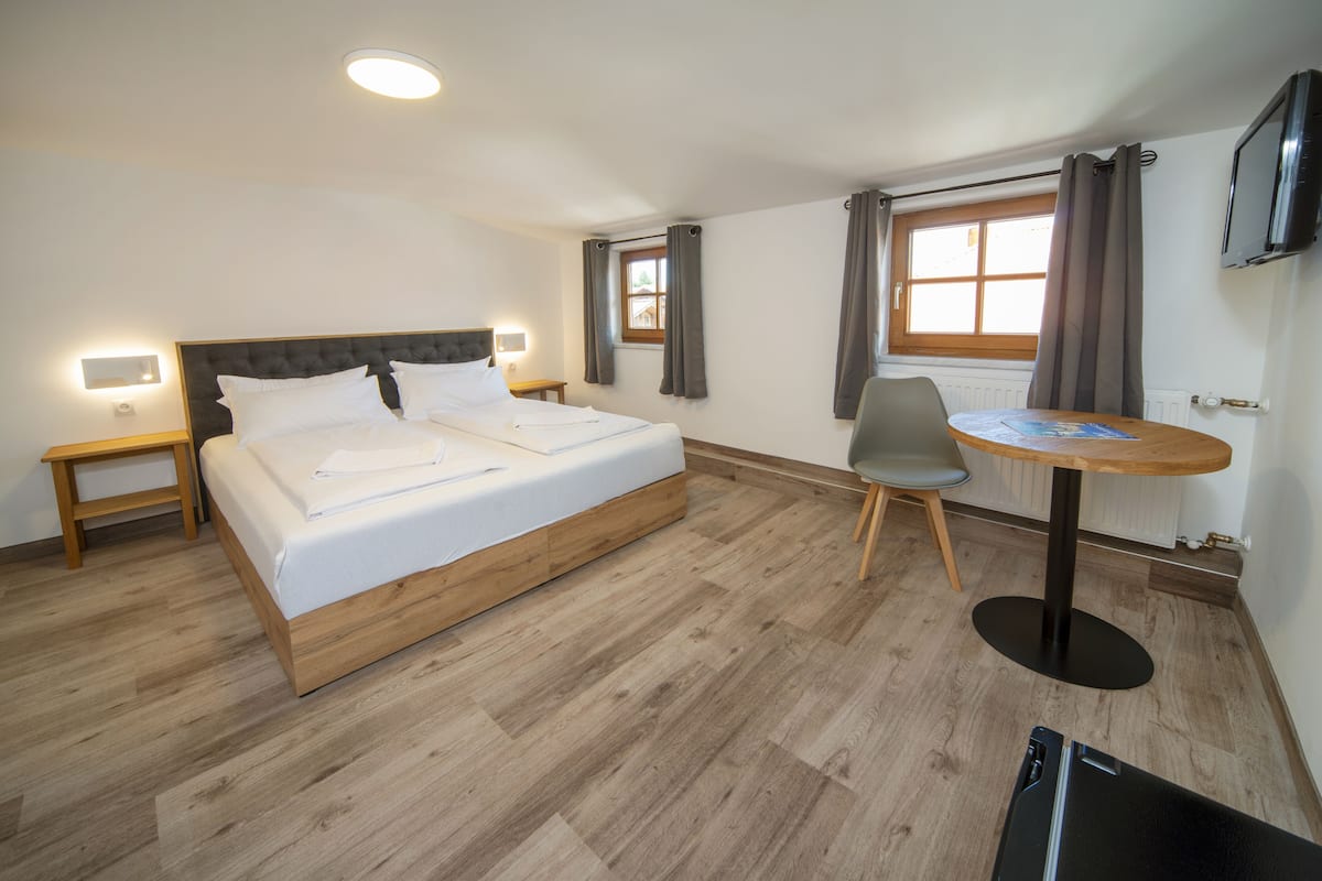 Ein Hotelzimmer mit einem hölzernen Bett, einem kleinen Tisch, einem Stuhl und einem an der Wand befestigten Fernseher.