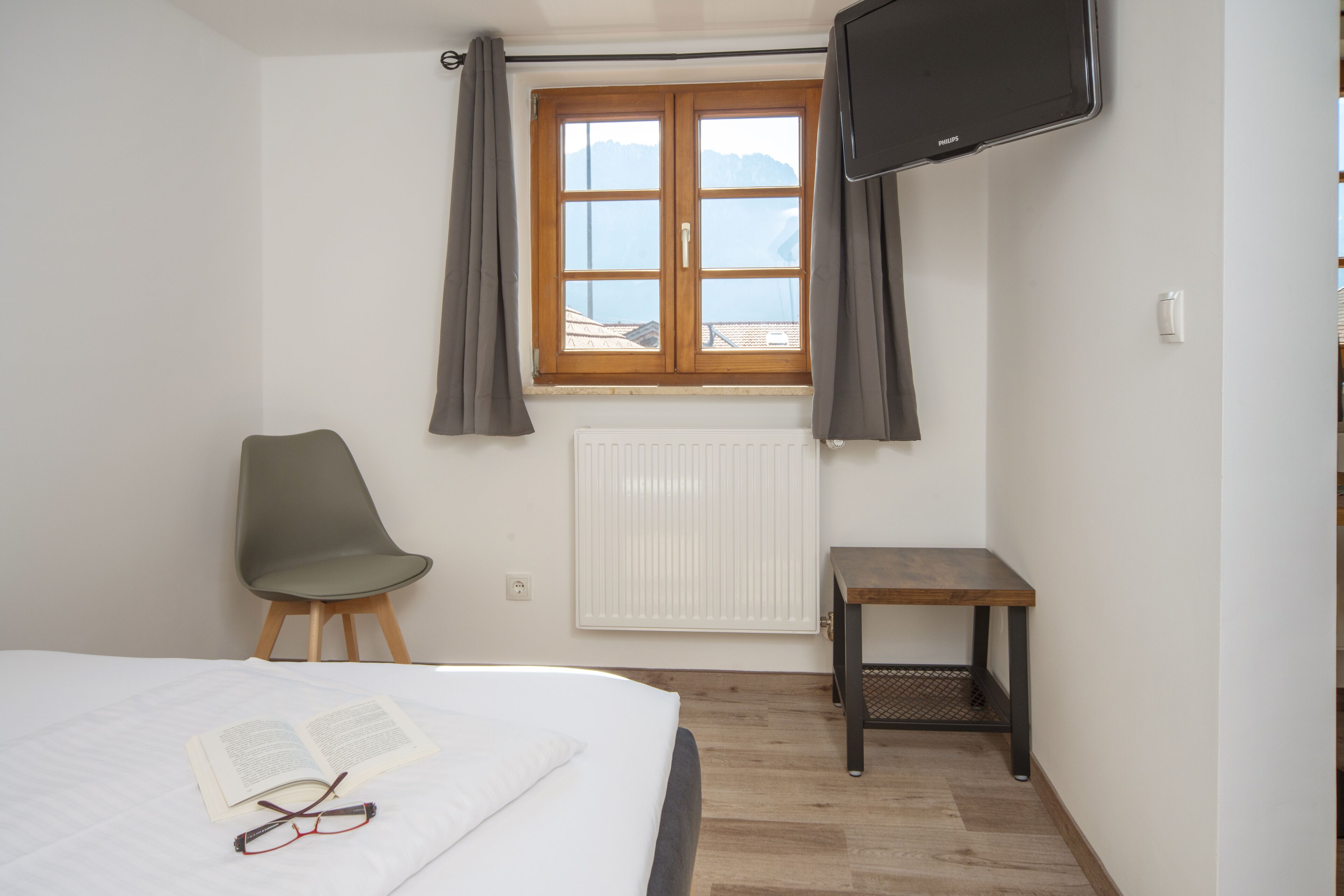 Un dormitorio con cama, un libro abierto, gafas, una silla, una mesita, una ventana con cortinas, un radiador y un televisor montado en la pared.