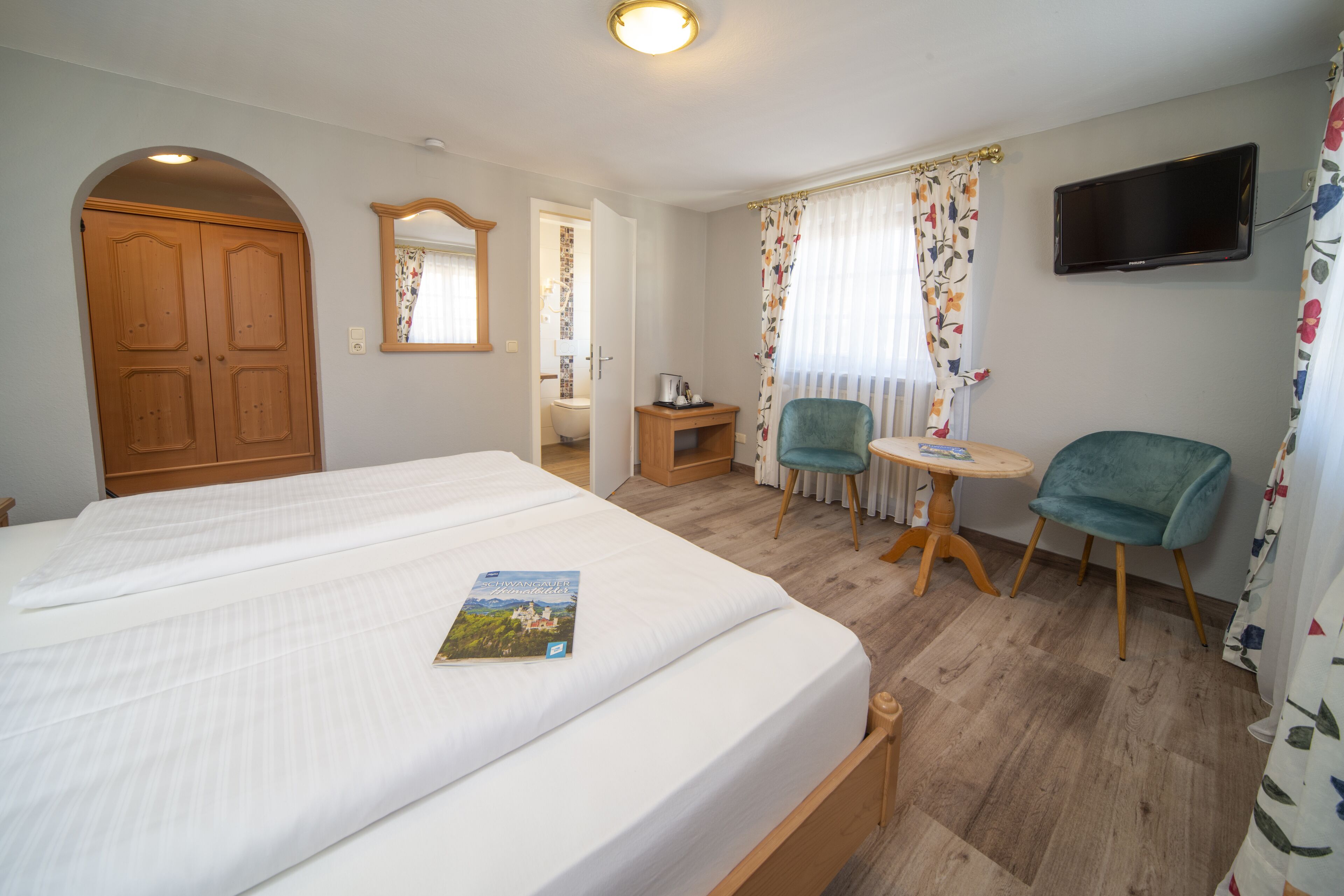 Ein Hotelzimmer mit einem großen Bett, einem kleinen Tisch mit Stuhl, einem an der Wand befestigten Fernseher und einer Holztür.