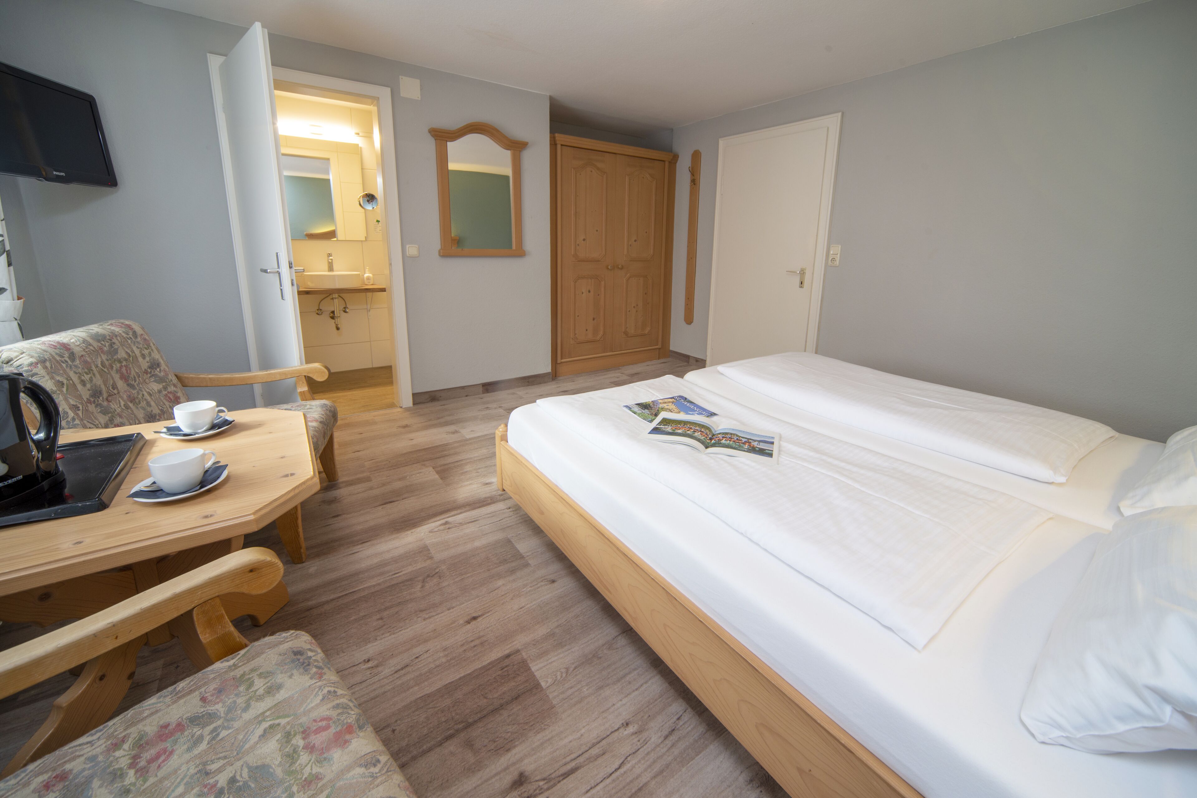 Ein Schlafzimmer mit einem Bett, einer hölzernen Bank, einem Tisch mit Tasse und Untertasse, einem Spiegel und einem Kleiderschrank.