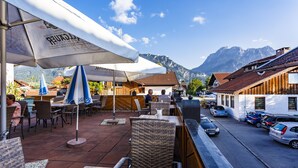Ein Café im Freien mit Korbstühlen und Sonnenschirmen, eine Straße mit parkenden Autos und einem Berg im Hintergrund.