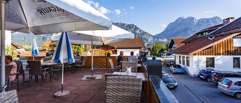 Een terras met rieten stoelen en parasols, een straat met geparkeerde auto's, en een berg op de achtergrond.