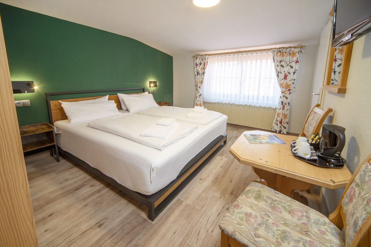 Ein Hotelzimmer mit einem großen Bett, einem hölzernen Schreibtisch mit Stuhl, einem kleinen Tisch mit Wasserkocher und einem Fenster mit blumigen Vorhängen.