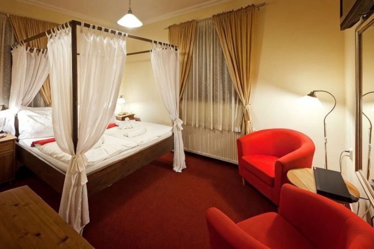 Una cama con dosel y cortinas, un sillón rojo, una mesita de noche y una lámpara.