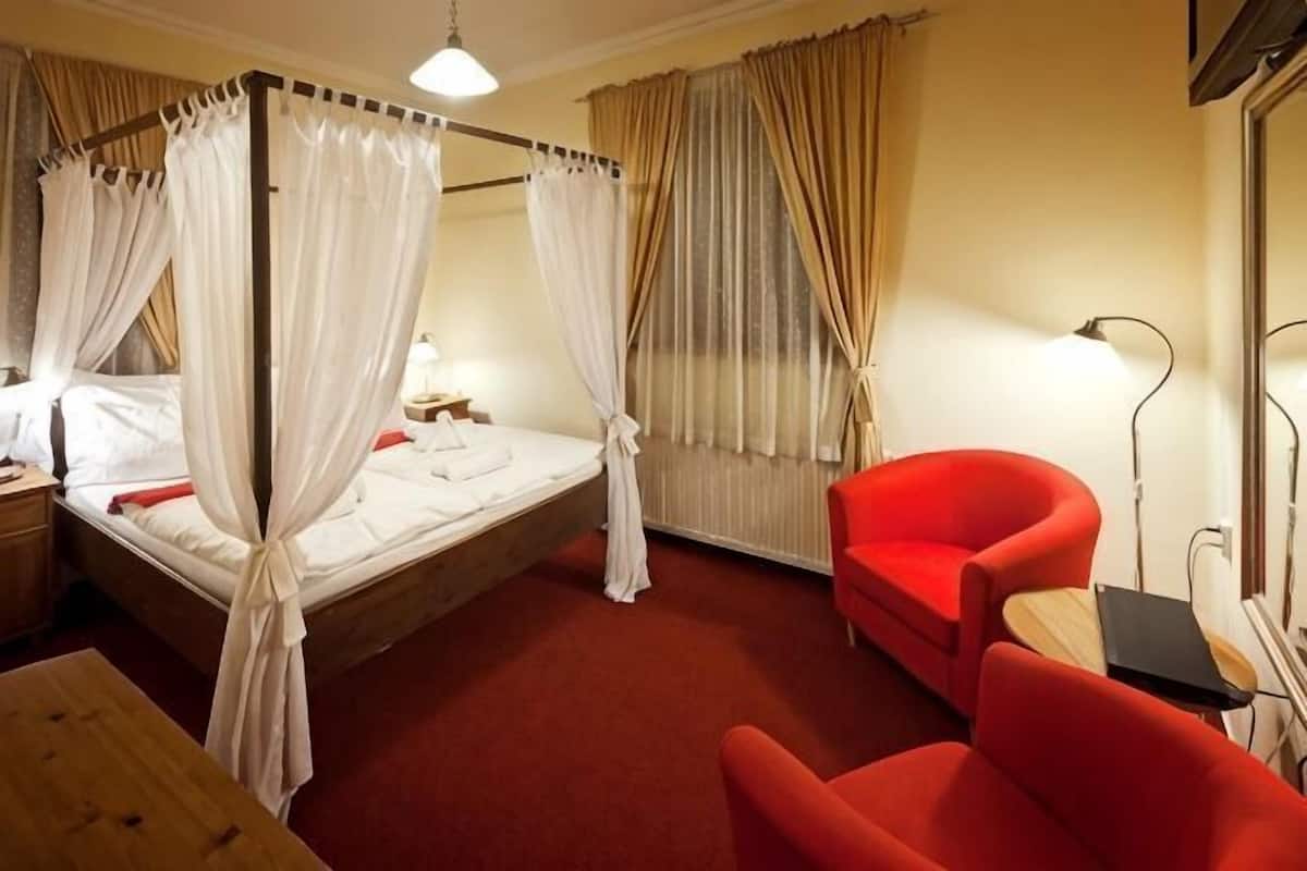 Una cama con dosel y cortinas, un sillón rojo, una mesita de noche y una lámpara.